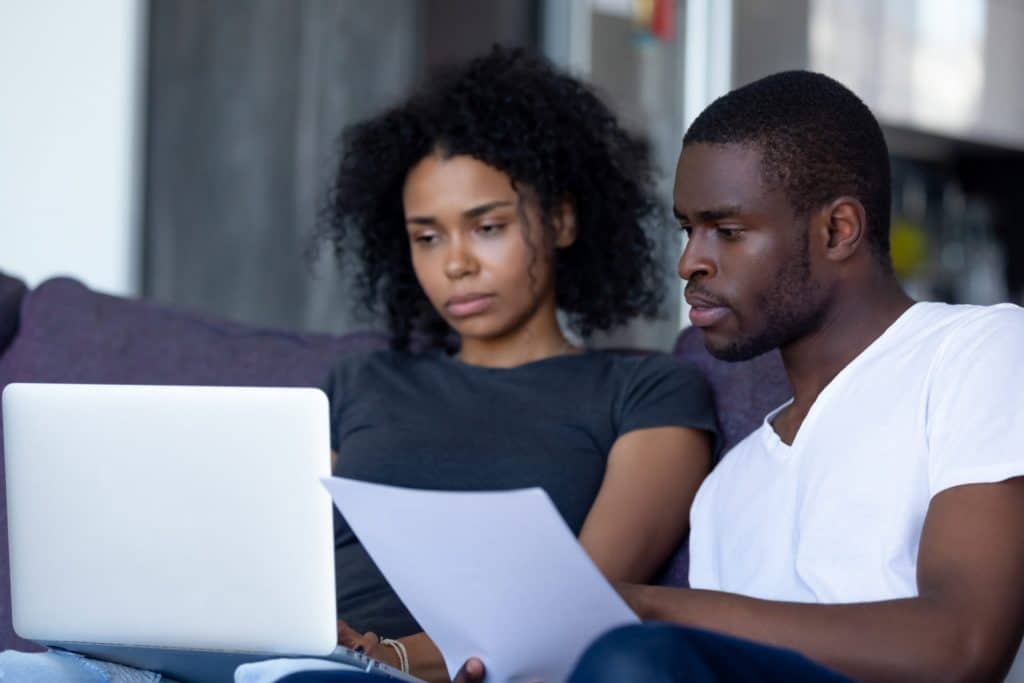 Serious african american couple reading document letter pay domestic bills checking bank account online on laptop at home, black family holding paper planning budget money expenses with computer app